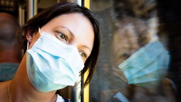 Woman wearing surgical mask leaning her head against window, looking outside