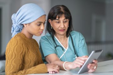A female cancer patient wearing a head wrap and her physician review medical documents on a tablet.