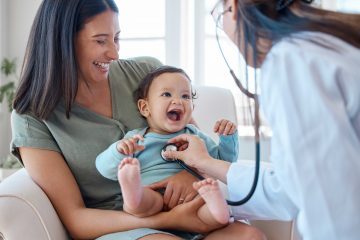 Shot of a baby sitting on her mother's lap while being examined by a doctor.