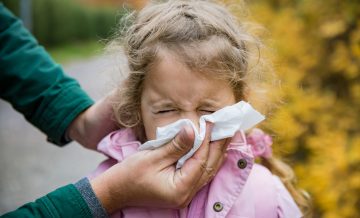 Father wiping daughter's nose with handkerchief. Sick little girl with cold and flu standing outdoors. Preschooler sneezing, coughing, having runny red nose. Autumn street background.