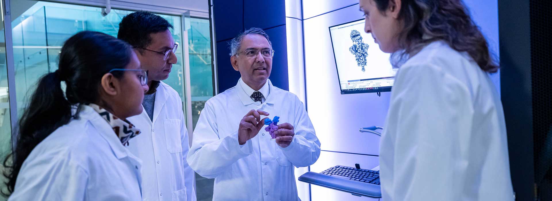 A photo of Dr. Sriram Subramaniam wearing a white lab coat and holding a 3D-printed atomic model of the COVID-19 Omicron variant spike protein. He is surrounded by three members of his lab wearing white lab coats, standing in front of a cryo-electron microscope and a computer screen.