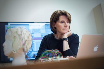 Dr. Lara Boyd sitting at a desk with a laptop, next to a mannequin head fitted with electrodes.