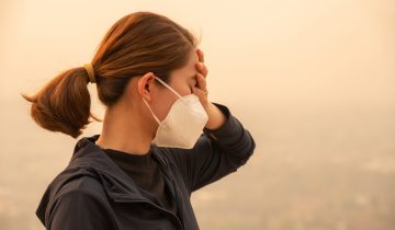 Person in a face mask stands outdoors against a hazy background.