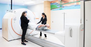 Two medical imaging technologists guide a patient through a PET/CT scan in a brightly lit imaging room at BC Cancer – Vancouver.