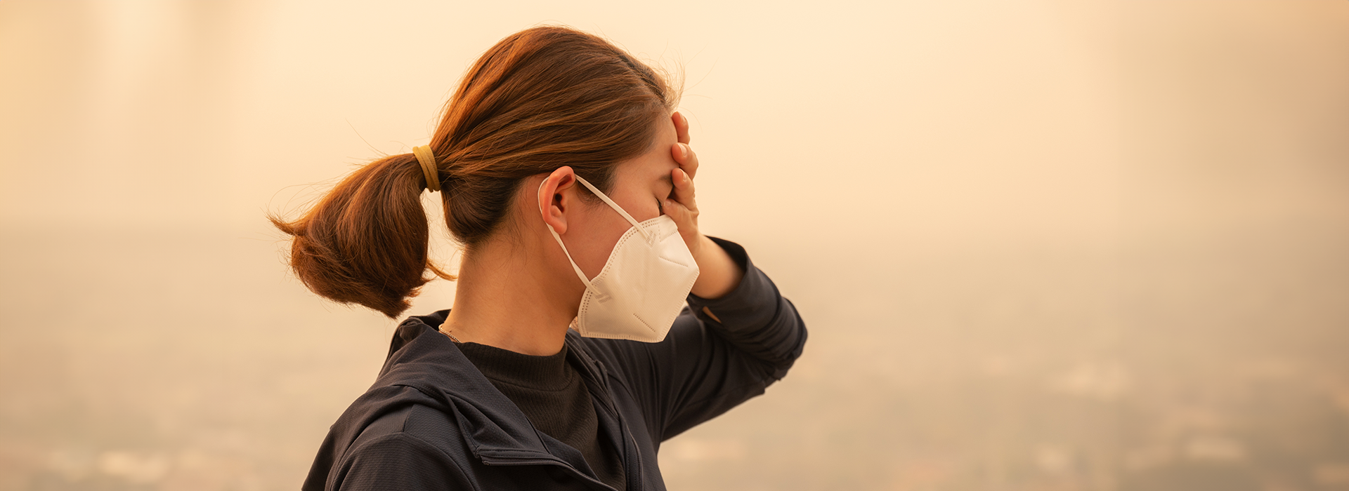 An individual with a mask on holds their hand to their face with a smokey city in the background.